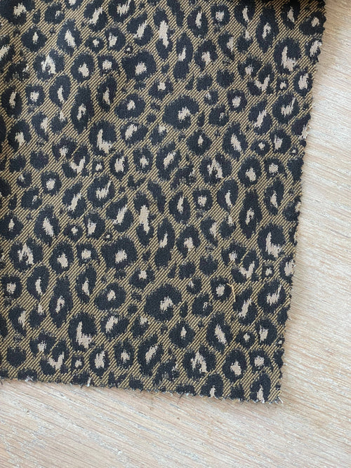 Amelia leopard jacquard fabric by Atelier 27, black and cream leopard spots woven into a warm brown ground, folded corner on pale wood